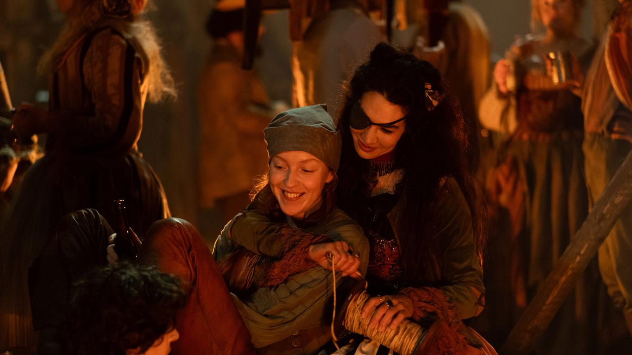 Raffaela und Rosa sitzen am Lagerfeuer, Rosa wickelt eine Schnur auf, hält Raffaela im Arm - beide lachen den gegenüber sitzenden Wolfgang Amadeus an