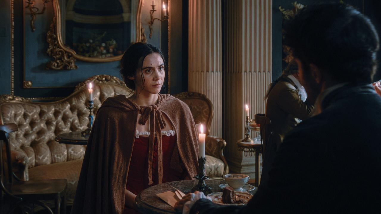 Maria Anna Mozart sitzt in rotem Kleid und braunem Umhang an einem kleinen Tisch in einem elegant eingerichteten Raum, gegenüber von Antonio Salieri