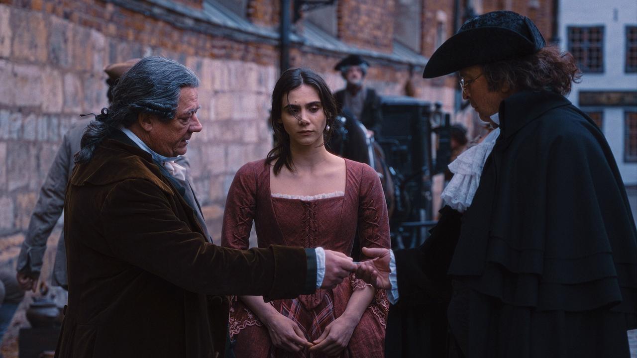 © ORF/WDR/Story House Pictures/Armands Virbulis Leopold Mozart und Dr. Wagner in historischer Kleidung stehen einander gegenüber und reichen sich die Hand, während Anna Maria in rotem Kleid zwischen ihnen steht