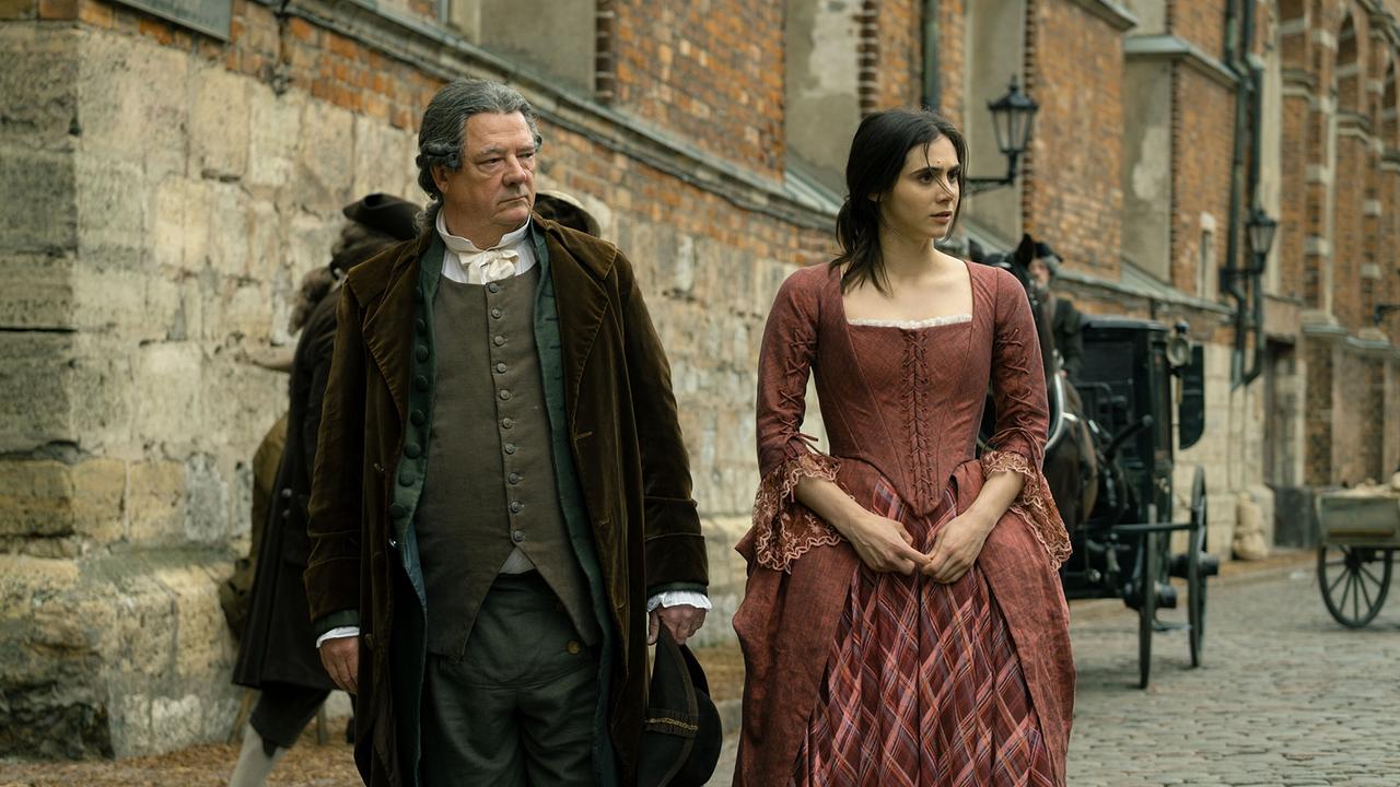 © ORF/WDR/Story House Pictures/Armands Virbulis Leopold Mozart geht mit seiner Tochter Maria Anna eine gepflasterte Straße entlang, hinter ihnen eine Kutsche und alte Backsteinmauern