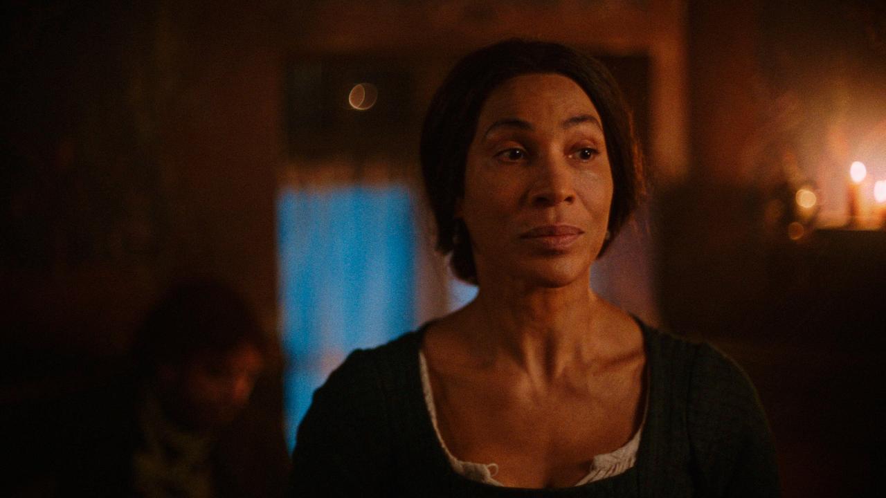 © ORF/WDR/Story House Pictures Eleonora steht in einem schwach mit Kerzenlicht beleuchteten Raum, im Hintergrund lässt sich eine Person schemenhaft im Schatten erkennen