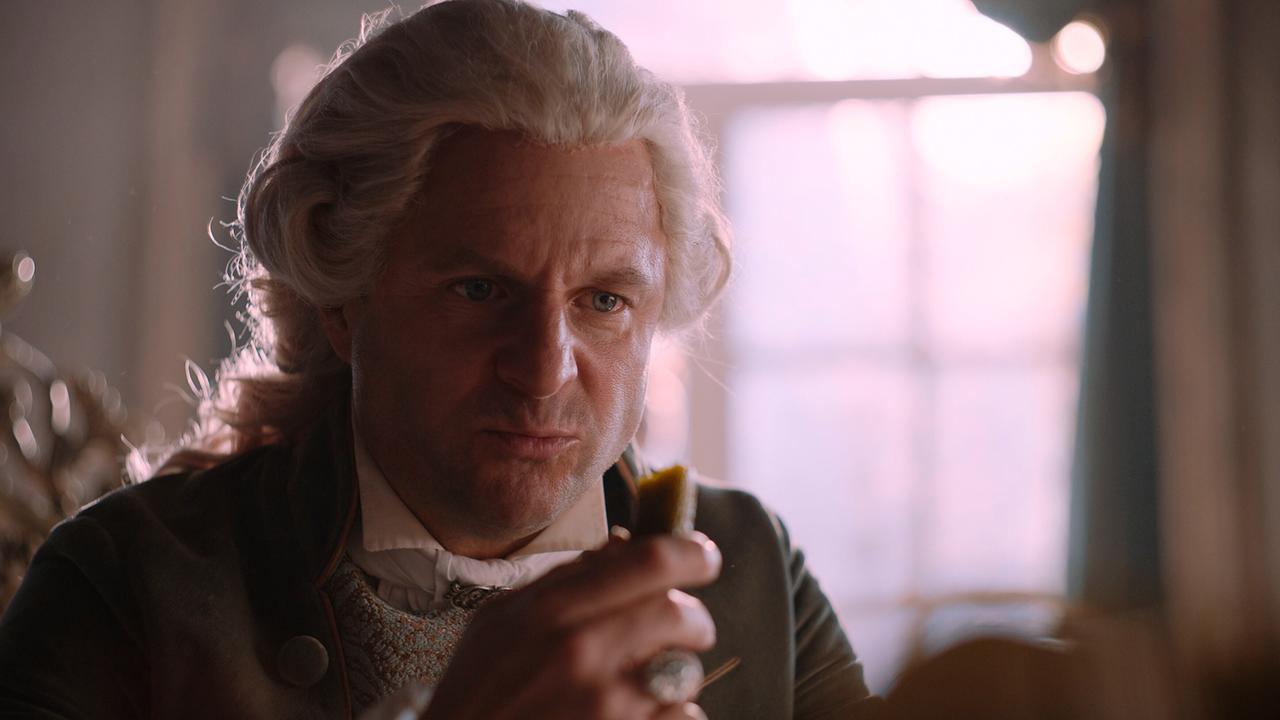 © ORF/WDR/Story House Pictures Philipp Hochmair als Kaiser Joseph II. mit gepuderter Perücke sitzt am Tisch und hat eine abgebissene Essiggurke in der Hand