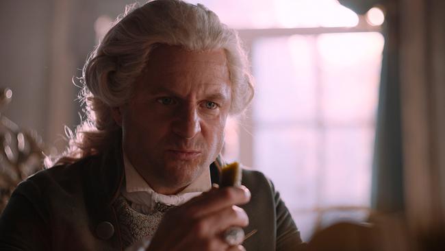 Philipp Hochmair als Kaiser Joseph II. mit gepuderter Perücke sitzt am Tisch und hat eine abgebissene Essiggurke in der Hand