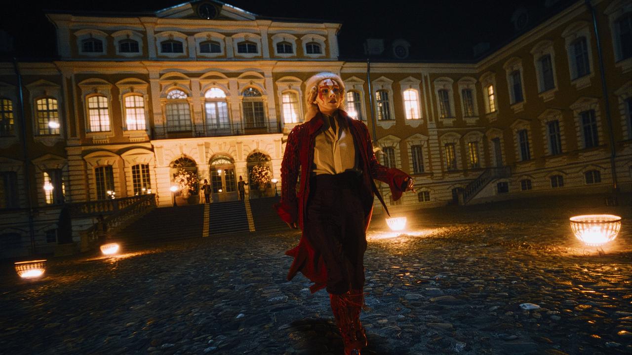 Eine weiß geschminkte Person in auffälliger roter historischer Kleidung geht nachts über einen Hof eines prunkvoll erleuchteten Palastes