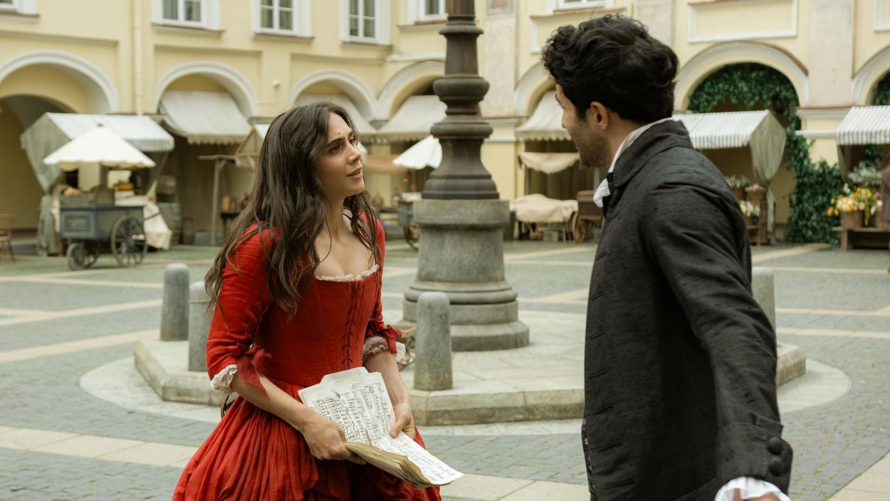 Anna Maria im roten Kleid hält Blätter der Partitur, auf einem Marktplatz steht ihr ihr Salieri gegenüber, mit dem sie sich unterhält
