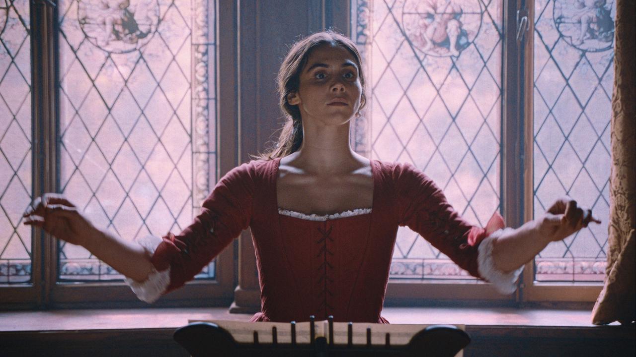 Maria Anna Mozart steht im roten Kleid am Dirigentenpult, sie steht vor großen historisch bemalten Fenstern 