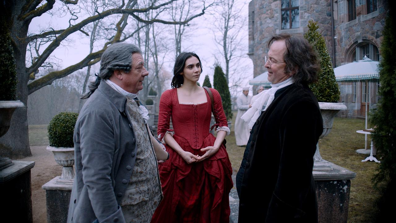 Leopold Mozart und Dr. Wagner stehen einander gegenüber, Maria Anna im roten Kleid zwischen ihnen - im Garten eines Schlosses an einem wolkenverhangenen, herbstlichen Tag