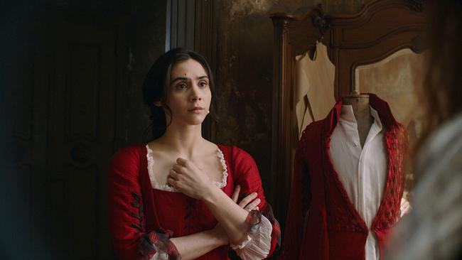 Maria Anna steht im roten Kleid in einem dunklen Raum, neben ihr eine Schneiderpuppe mit weißen Hemd und roten Mantel