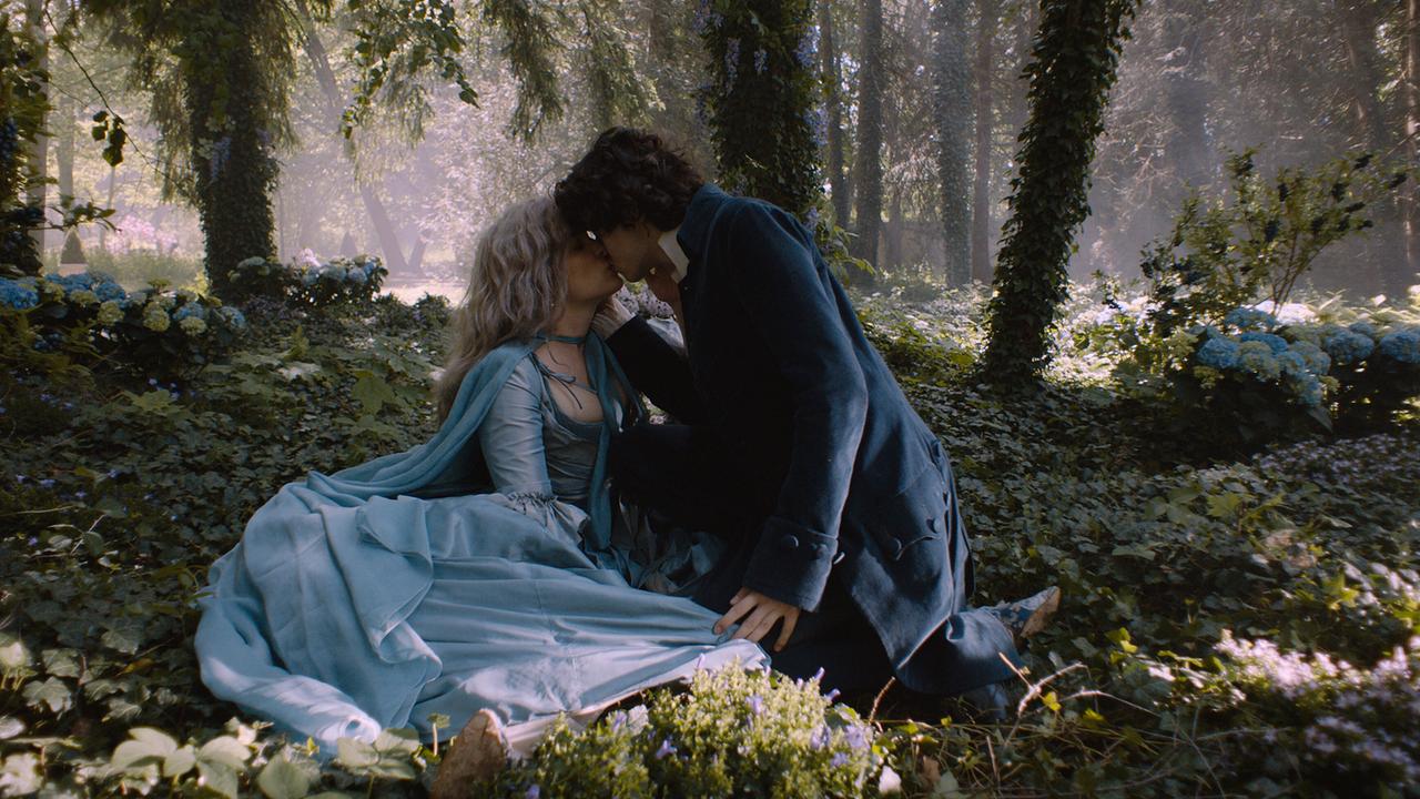 Wolfgang Amadeus und Marie Antoinette, beide in eleganter historischer Kleidung, sitzen in einem sonnendurchfluteten Wald und küssen sich zärtlich