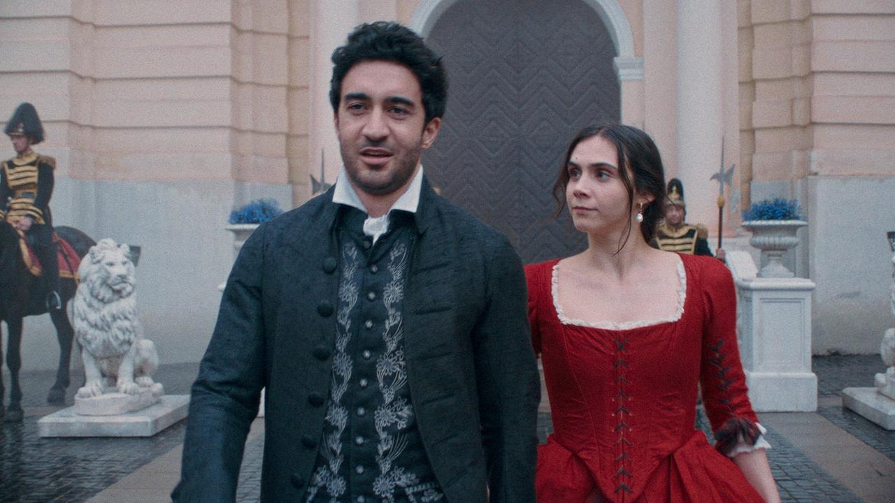 Antonio Salieri in dunkler historischer Kleidung und Maria Anna im roten Kleid gehen nebeneinander vor einem Schlossportal, flankiert von Wachen und steinernen Löwen