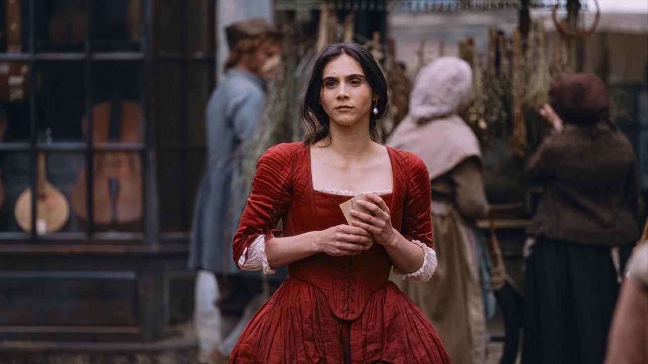 © ORF/WDR/Story House Pictures/Armands Virbulis Maria Anna Mozart im roten Kleid, vor einem Musikaliengeschäft auf der Straße stehend, hält sie Zettel in der Hand und blickt in die Ferne