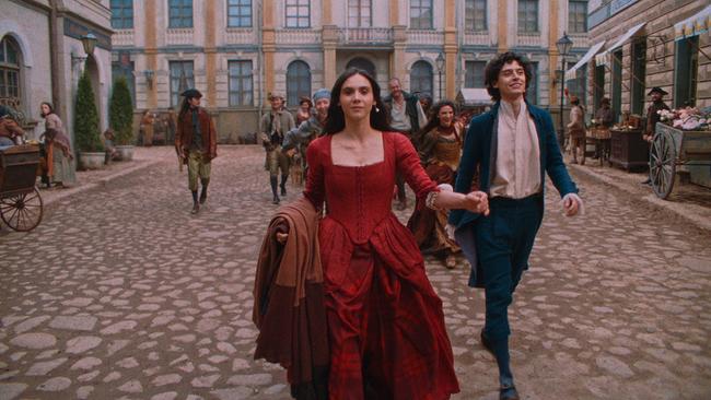 Anna Maria im roten Kleid und Wolfgang Amadeus im blauen Anzug laufen lächelnd eine gepflasterte Straße entlang, ihnen folgt eine Gruppe einfach gekleideter Personen