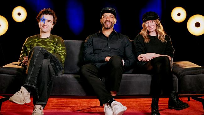 COSMÓ, Cesár Sampson und Zoe Straub sitzen nebeneinander auf einem Sofa in einem Studio mit dunklem Hintergrund und runden Lichtquellen.