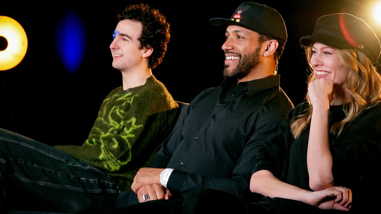 Seitenansicht von COSMÓ, Cesár Sampson und Zoe Straub nebeneinander auf einem Sofa in einem Studio mit dunklem Hintergrund und runden Lichtquellen
