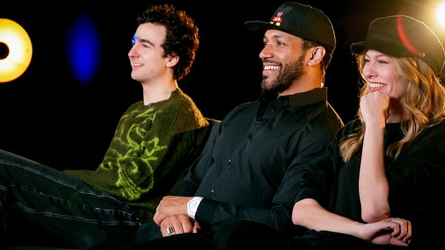 Seitenansicht von COSMÓ, Cesár Sampson und Zoe Straub nebeneinander auf einem Sofa in einem Studio mit dunklem Hintergrund und runden Lichtquellen