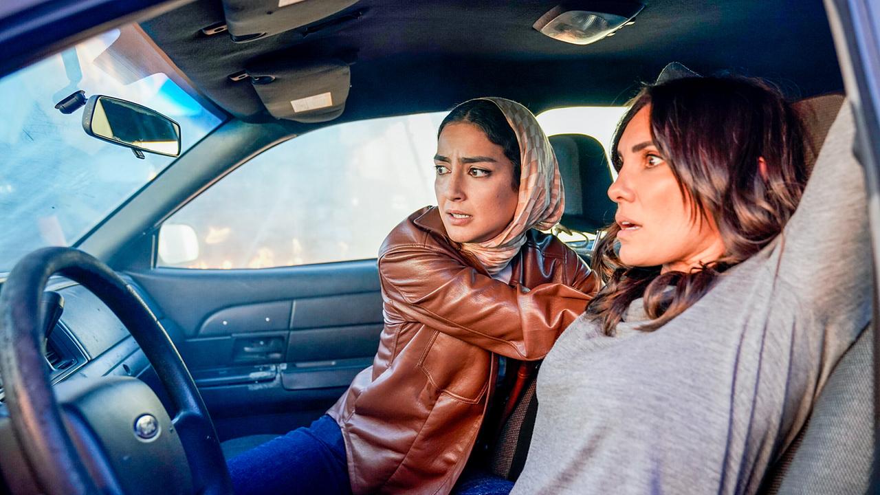 Fatima und Kensi sitzen im Auto, Fatima sitzt am Beifahrersitz und holt etwas von hinten, Kensi hält die Hände nach oben, beide Frauen blicken gebannt nach vorne