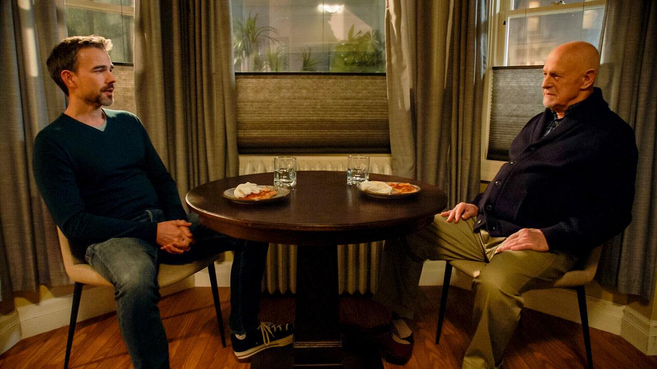 Ein junger und ein älterer Mann sitzen sich an einem Wohnzimmertisch gegenüber, auf dem Tisch Gläser und Teller mit Pizzastücken