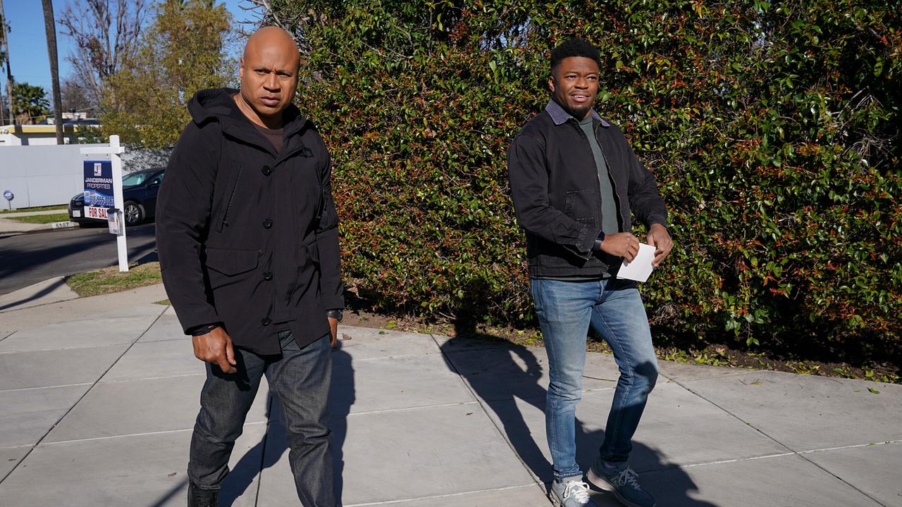 Zwei Männer in Jeans und schwarzen Jacken gehen den gepflasterte Einfahrt eines Hauses entlang, hinter ihnen eine hohe Gartenhecke