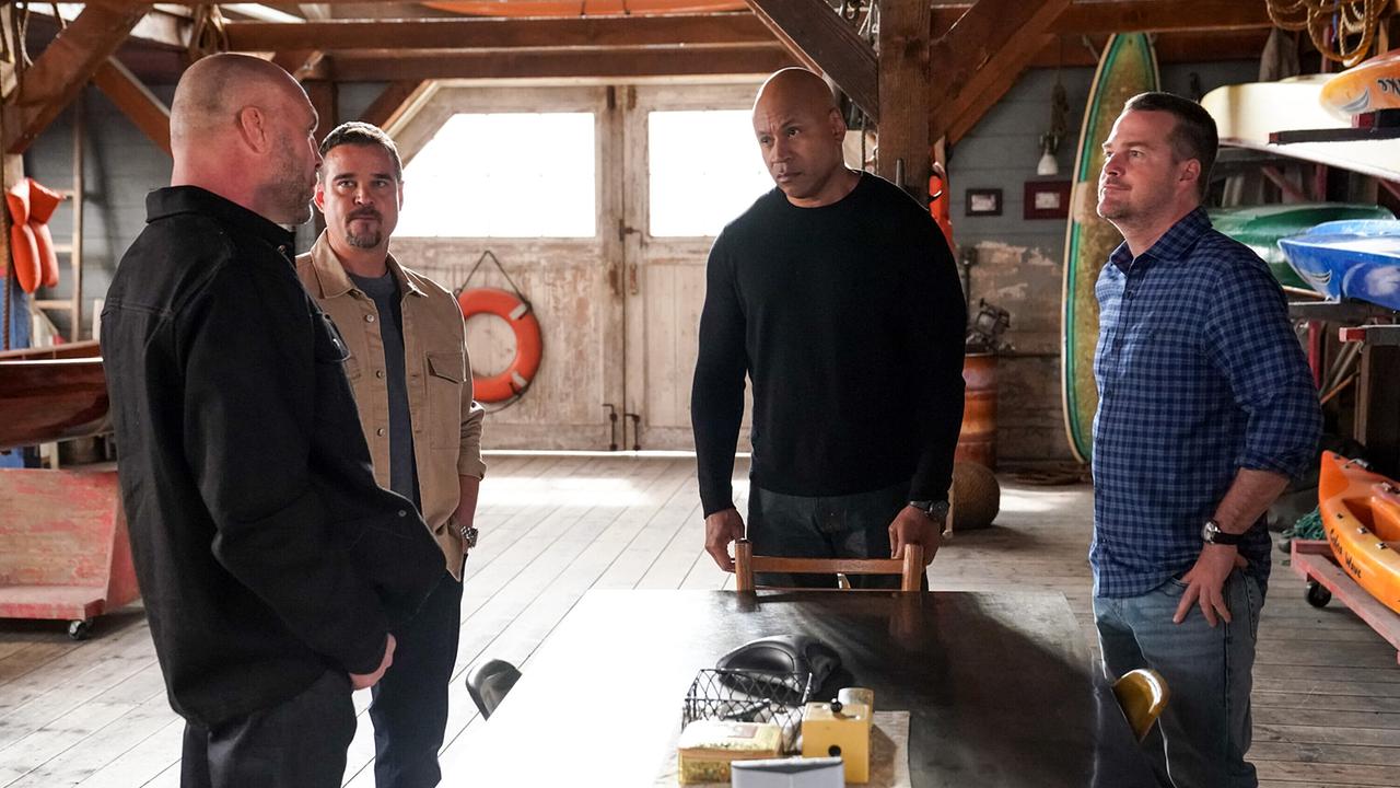 Vier Männer stehen in einem holzvertäfelten Raum mit Bootsausrüstung und Surfbrettern, offenbar im Gespräch um einen Tisch