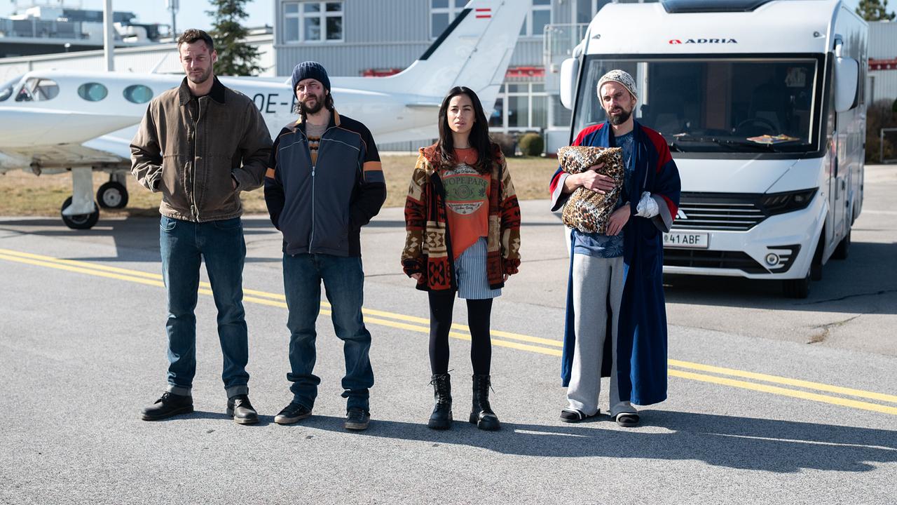 Flo Kienzl, Eddi Kovac, Samira Leoni und Jan Miracek stehen vor einem Wohnmobil und einem Kleinflugzeug auf einem Flugplatz.
