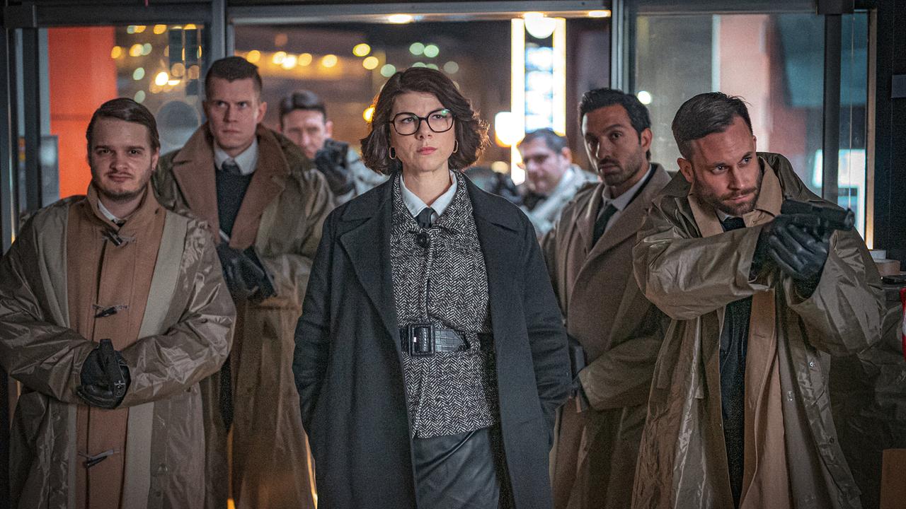 Sybille Danninger mit Brille geht mit einer Gruppe von mit Pistolen bewaffneten Männern in Mänteln durch einen nächtlich beleuchteten Eingang.