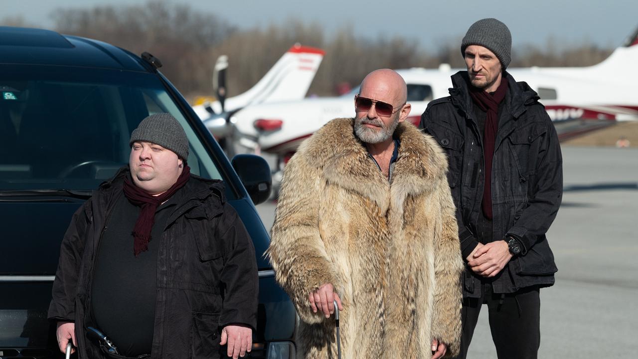 Nogger im Pelzmantel und mit Sonnenbrille steht mit Brain und Pinky, beide schwarz gekleidet und mit Haube,  auf einem Flugplatz vor einem Privatflugzeug.