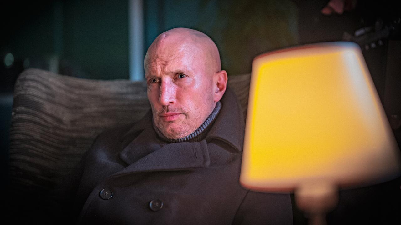Der glatzköpfige Thilo Mannheim sitzt mit ernster Miene  neben einer warm leuchtenden Tischlampe auf einem Sofa.