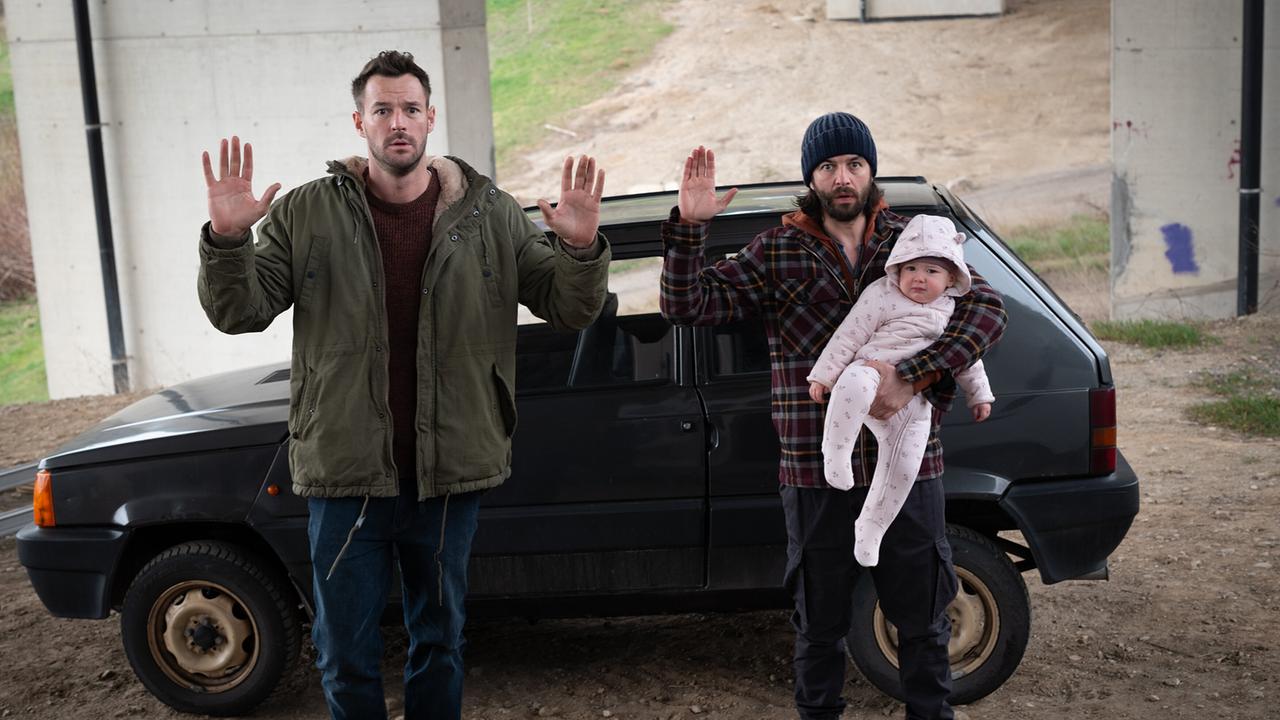 Flo Kienzl und Eddi Kovac, der ein Baby in einem Arm hält, ergeben sich vor einem Auto unter einer Brücke.