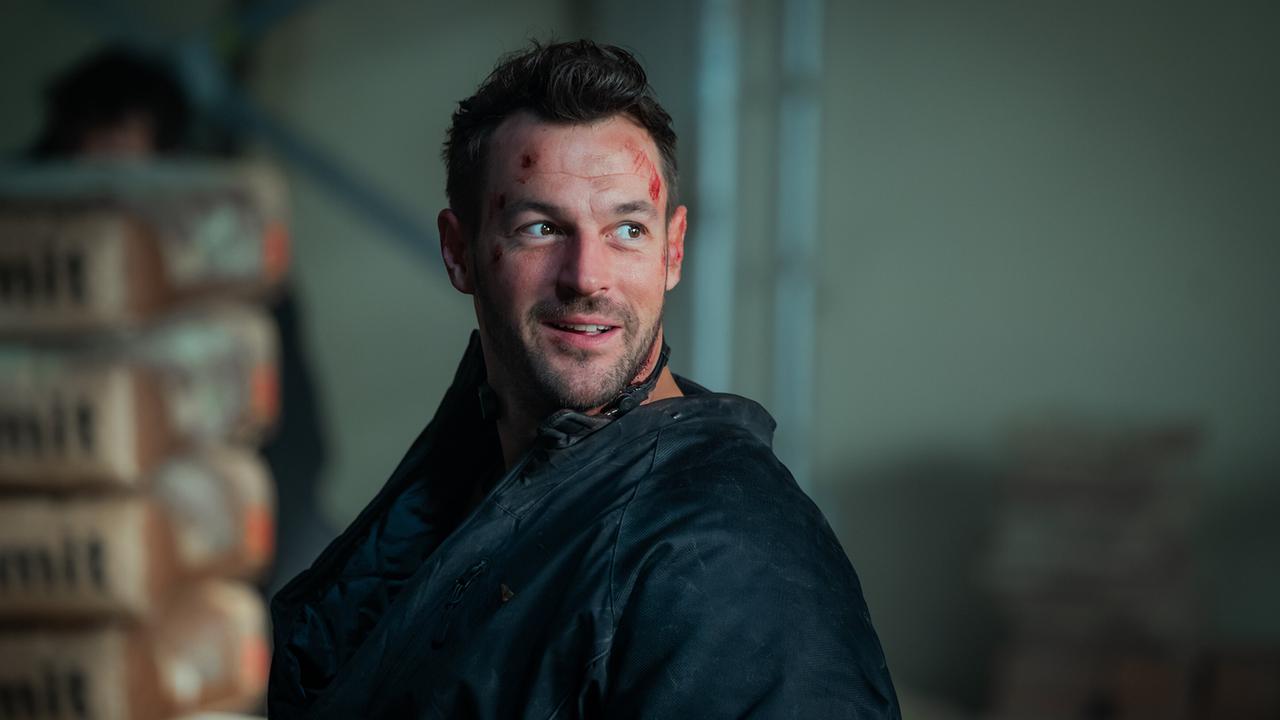 Der im Gesicht verletzte Flo Kienzl  in dunkler Jacke blickt mit einem unsicheren Lächeln zur Seite in einem dunklen Raum.