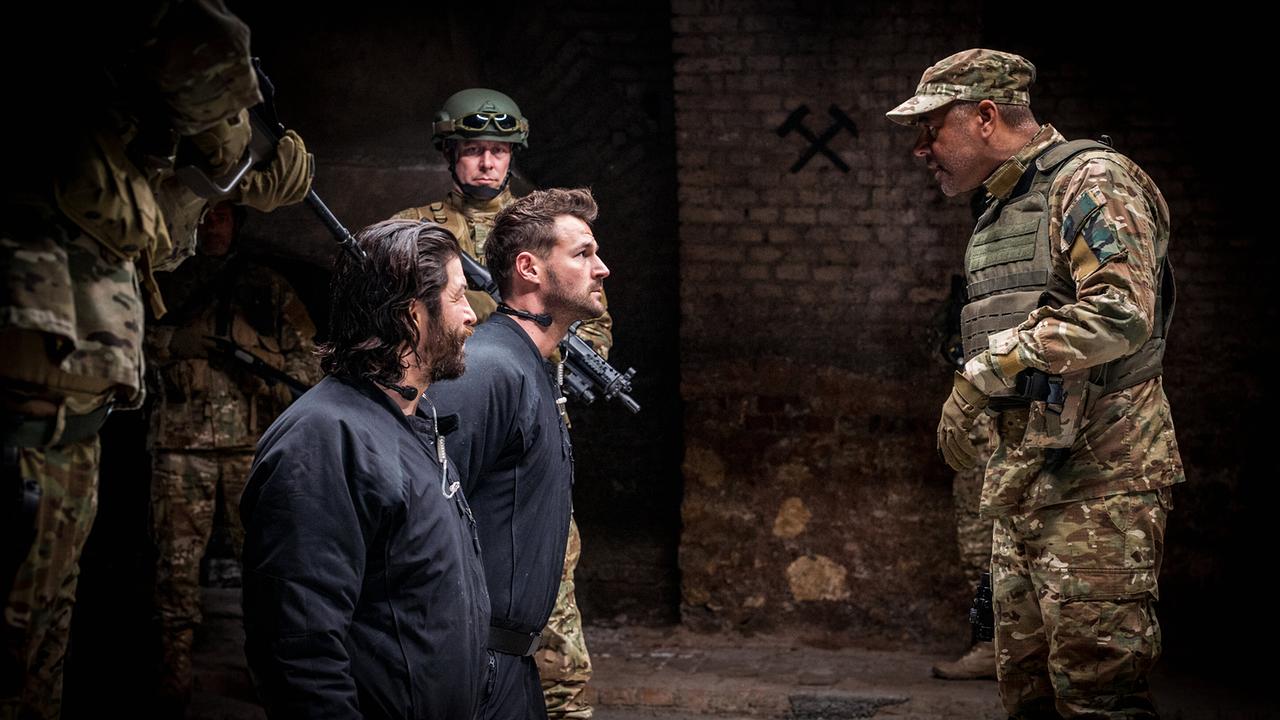 Flo Kienzl und Eddi Kovac in dunklen Overalls knien vor bewaffneten Soldaten in einem düsteren Raum.