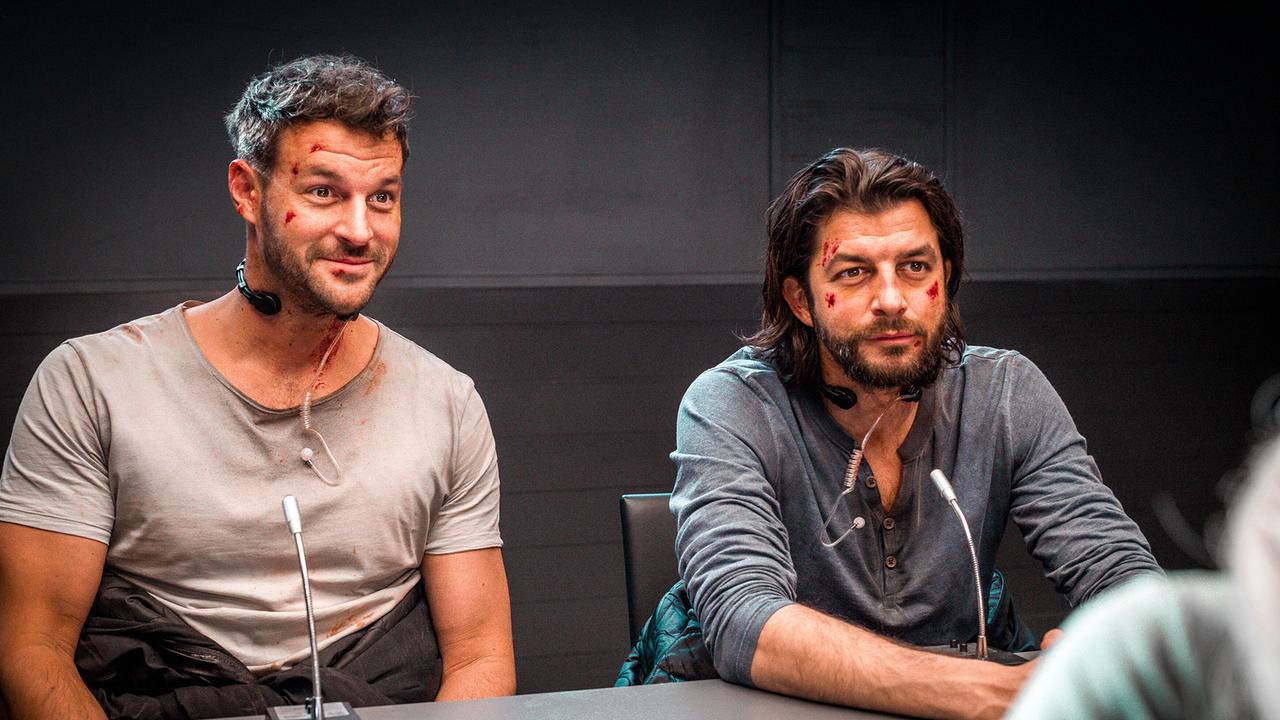 Flo Kienzl und Eddi Kovac mit Schürfwunden im Gesicht sitzen vor Mikrofonen an einem Tisch in einem dunklen Raum.