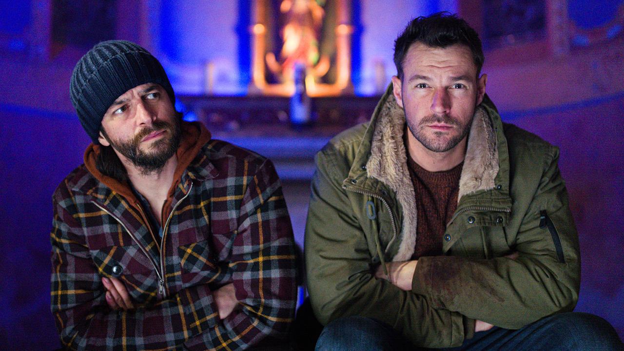 Eddi Kovac und Flo Kienzl sitzen mit verschränkten Armen in blau-violettem Licht vor einem religiösen Altar.