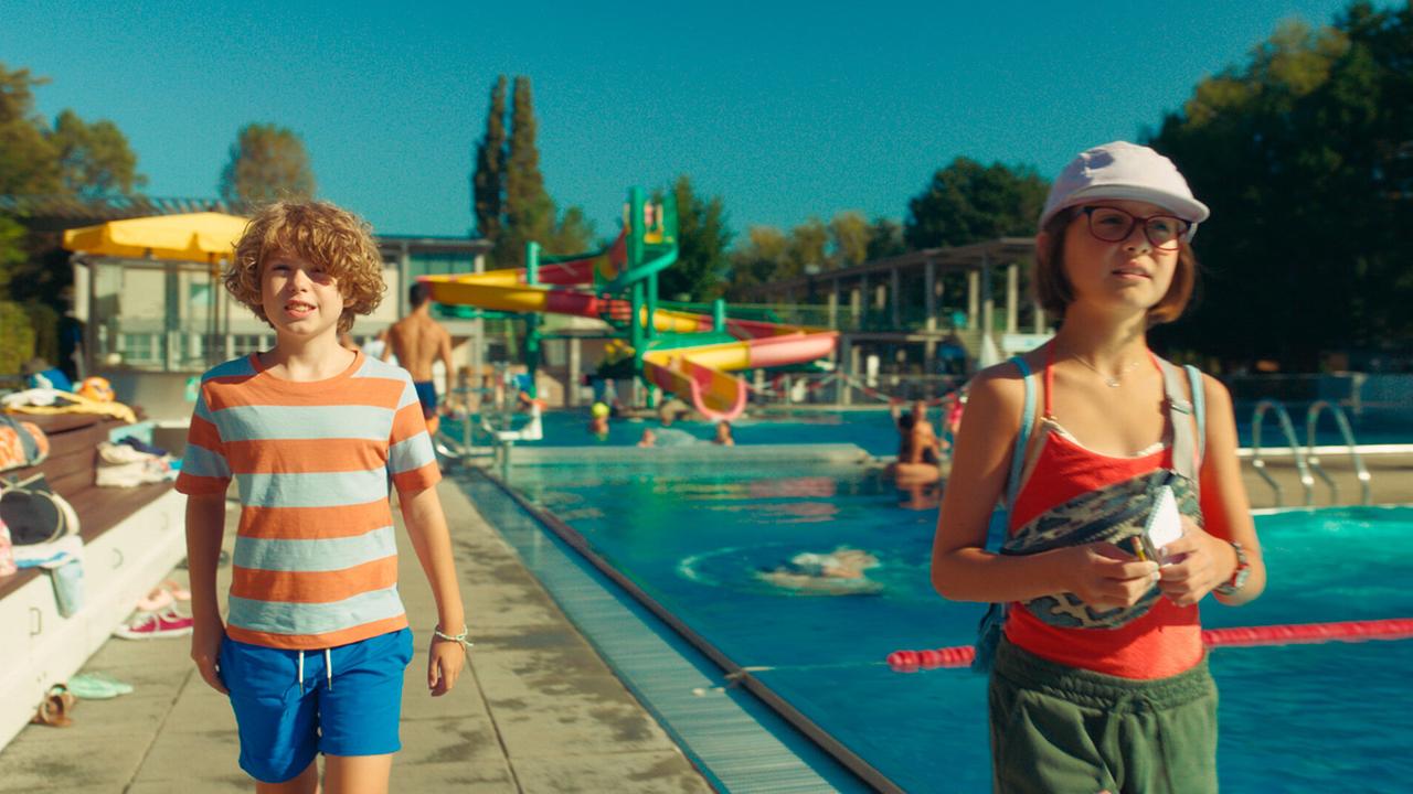 Franz und Gabi gehen nebeneinander an einem Freibadbecken entlang.