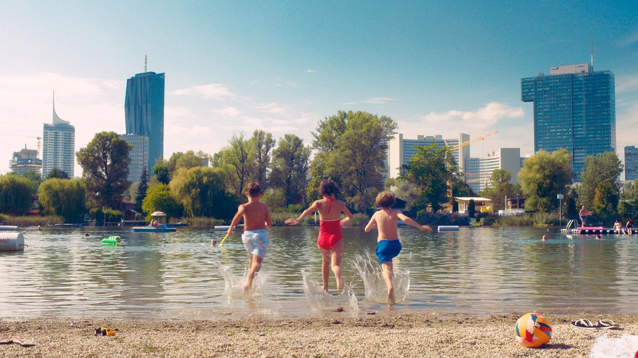 Eberhard, Gabi und Franz laufen ins Wasser eines Badesees mit Stadtsilhouette im Hintergrund.