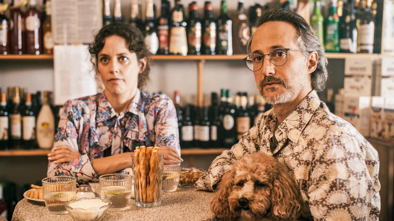 Vicky und Herr Bahram sitzen zusammen bei Tisch und blicken verwundert.