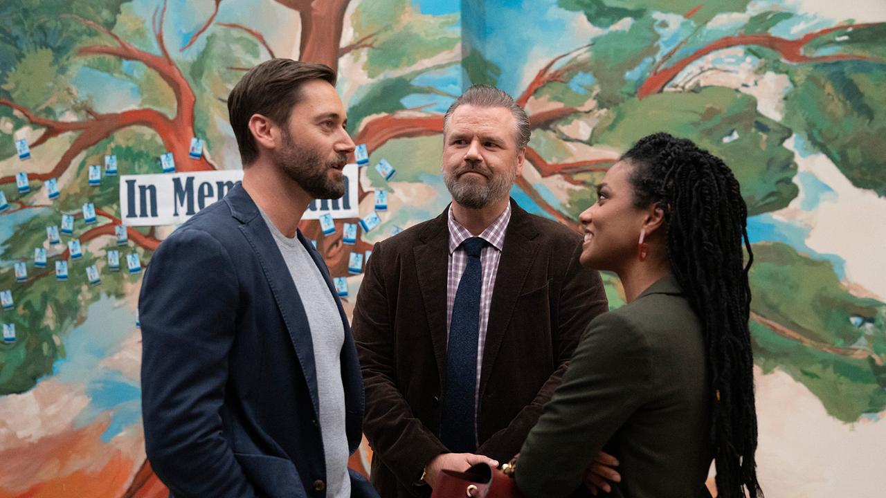 © ORF/Universal/NBC/Virginia Sherwood Eine Frau steht mit zwei Männern im Gespräch vor einer mit einem Baum bemalten Wand, auf den Ästen kleben Notizzettel um ein Schild mit der Aufschrift 'IN MEMORIAM'