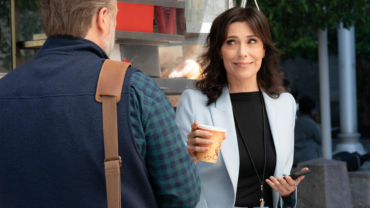 Eine Frau in hellblauem Hosenanzug und schwarzem Pulli hält einen Coffee-To-Go-Becher in der Hand und lächelt einen bärtigen, älteren Mann an, der ihr gegenübersteht