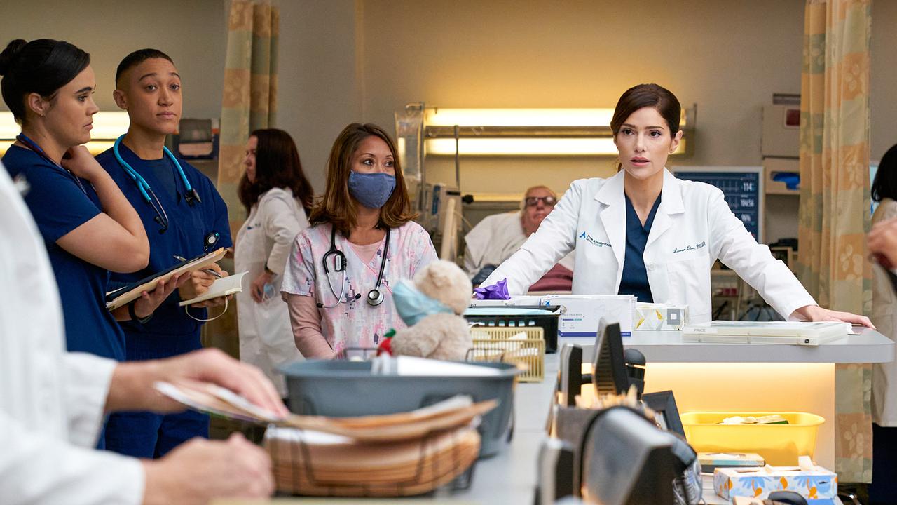 Mehrere Personen in medizinischer Kleidung stehen und sprechen in einem Krankenhaus, umgeben von medizinischen Geräten und Unterlagen