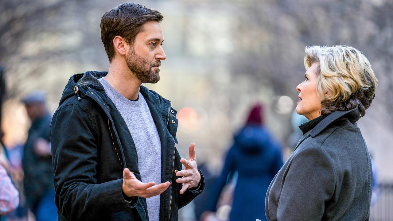 Eine winterliche Straßenszene im Freien - ein bärtiger Mann im blauen Pullover und dunklem Parka unterhält sich mit einer elegant gekleideten Frau im grauen Mantel