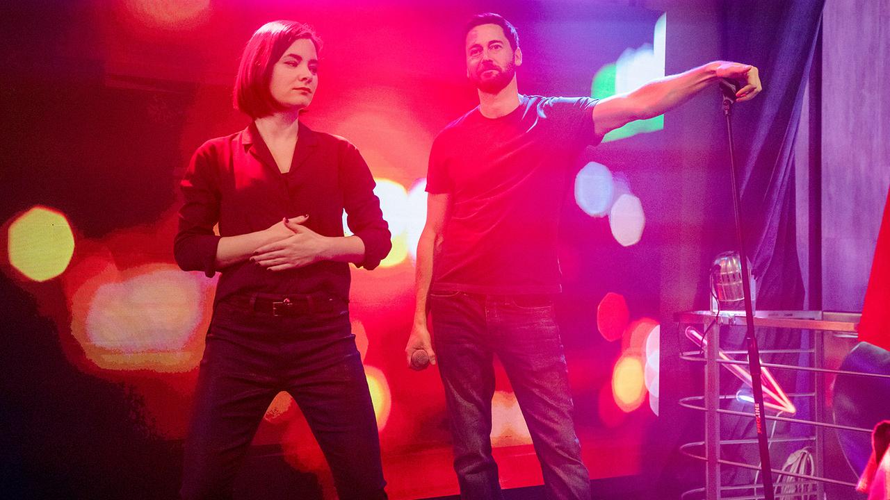 Eine Frau in schwarzer Bluse und schwarzer Hose und ein Mann in blauem T-Shirt und Blue Jeans stehen gemeinsam auf einer bunt beleuchteten Karaoke-Bühne