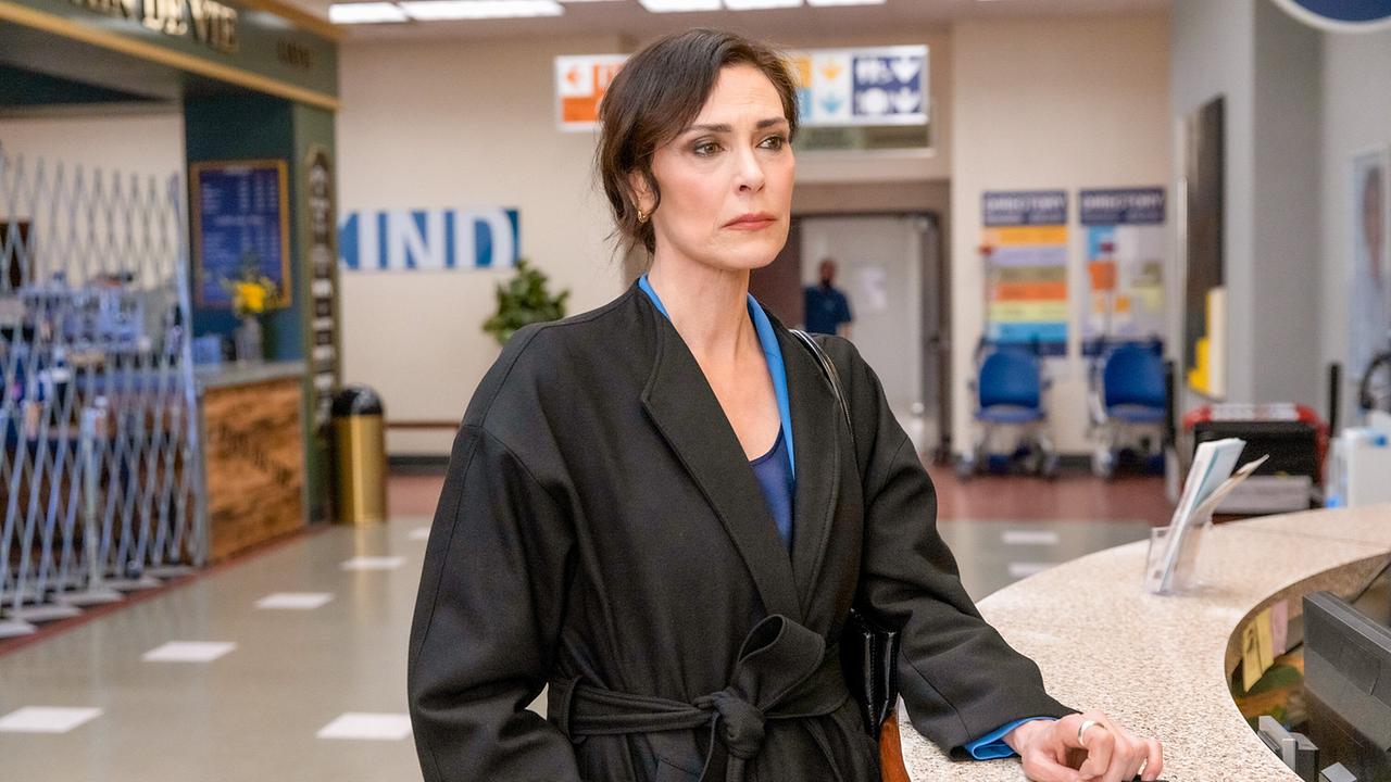 Dr. Veronica Fuentes in einem schwarzen Mantel und blauem Hosenanzug, sie steht bei der Aufnahme des Krankenhauses und legt mit ernstem Blick einen Ausweis auf die Theke