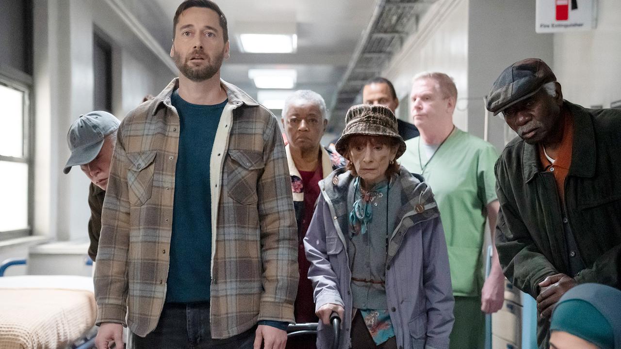 Im Gang eines Krankenhauses steht Dr. Max Goodwin in blauem Shirt und karierter Jacke, neben und hinter ihm stehen weißhaarige, ältere Frauen und Männer