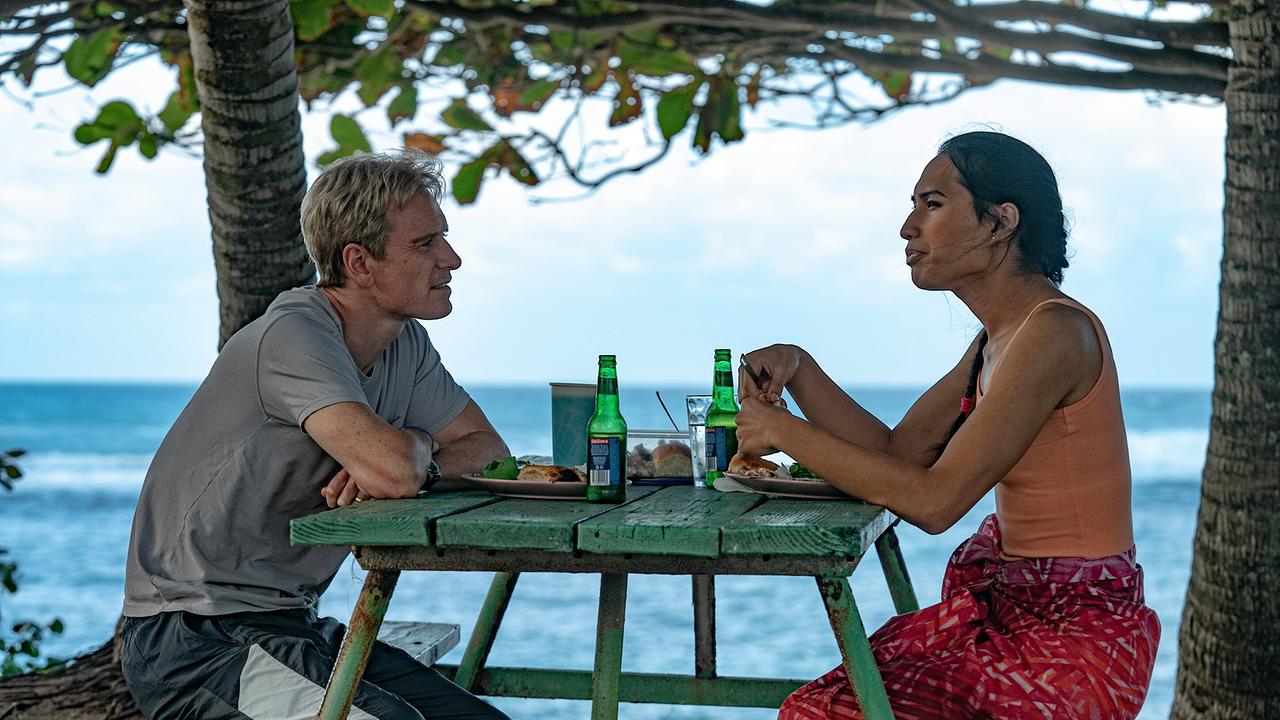 An einem schattigen Platz unter Bäumen sitzen Thomas Rongen und Jaiyah an einem Tisch und nehmen einen Snack zu sich, im Hintergrund paradiesisches Meer