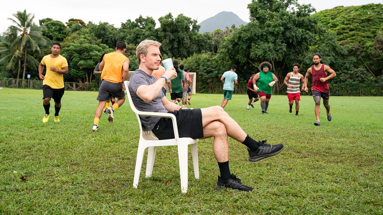 Michael Fassbender sitzt als Trainer Thomas Rongen mit einem Coffee-To-Go-Becher auf einem Fußballplatz, Männer laufen beim Training um ihn herum