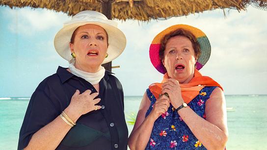 Lilibet und Omama mit Sonnenhut schauen geschockt am Strand auf etwas, im Hintergrund das Meer.