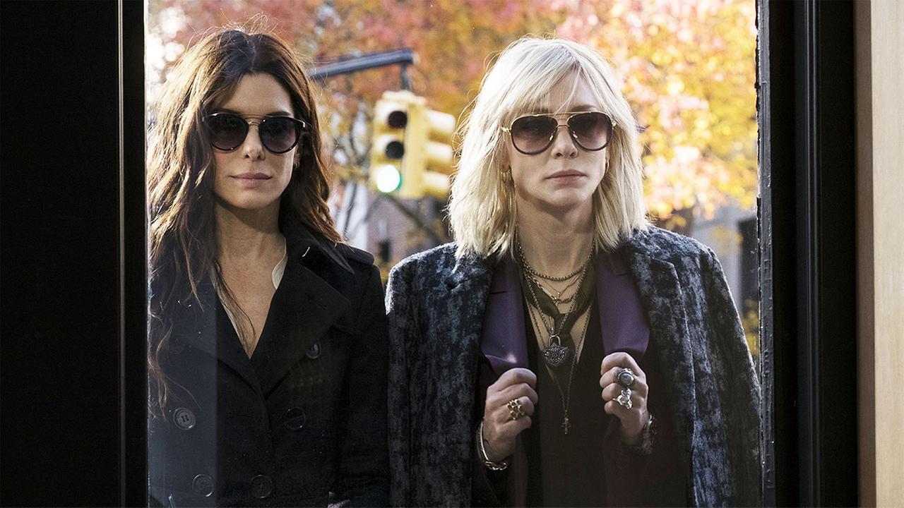© ORF/Pro7/© 2018 Warner Bros Sandra Bullock als Debbie Ocean und Cate Blanchett als Lou, dunkel bekleidet stehen die beiden mit Sonnenbrillen auf einer Straße, blicken in die geöffnete Türe eines Gebäudes und sind dabei den Raum zu betreten, im Hintergrund eine Ampel, die grün zeigt und herbstliche Bäume