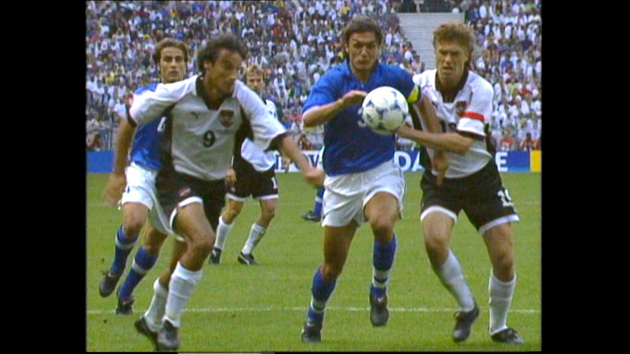 WM 1998 in Frankreich, die letzte WM mit österreichischer Beteiligung: hitzige Fußballszene, in der mehrere Spieler aus zwei Teams, unter ihnen Toni Polster, Ivica Vastic, um den Ball kämpfen, während im Hintergrund ein vollbesetztes Stadion zu sehen ist.