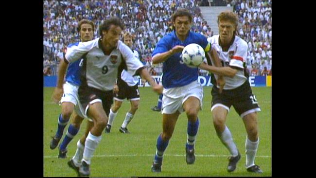WM 1998 in Frankreich, die letzte WM mit österreichischer Beteiligung: hitzige Fußballszene, in der mehrere Spieler aus zwei Teams, unter ihnen Toni Polster, Ivica Vastic, um den Ball kämpfen, während im Hintergrund ein vollbesetztes Stadion zu sehen ist.