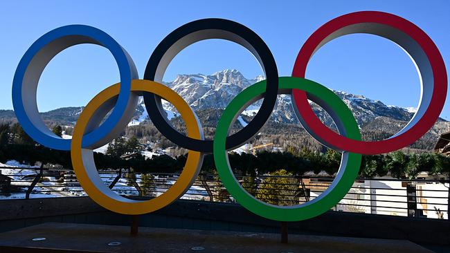 Die fünf olympischen Ringe stehen im Vordergrund, dahinter erstreckt sich eine sonnige Winterlandschaft in Cortina d'Ampezzo mit schneebedeckten Bergen.