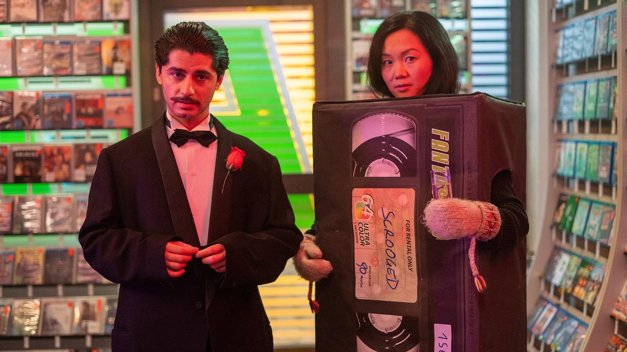 Enis im Smoking mit Masche und Rose am Revers steht neben Domino, die ein übergroßes VHS-Kassettenkostüm trägt, in einer Videothek.